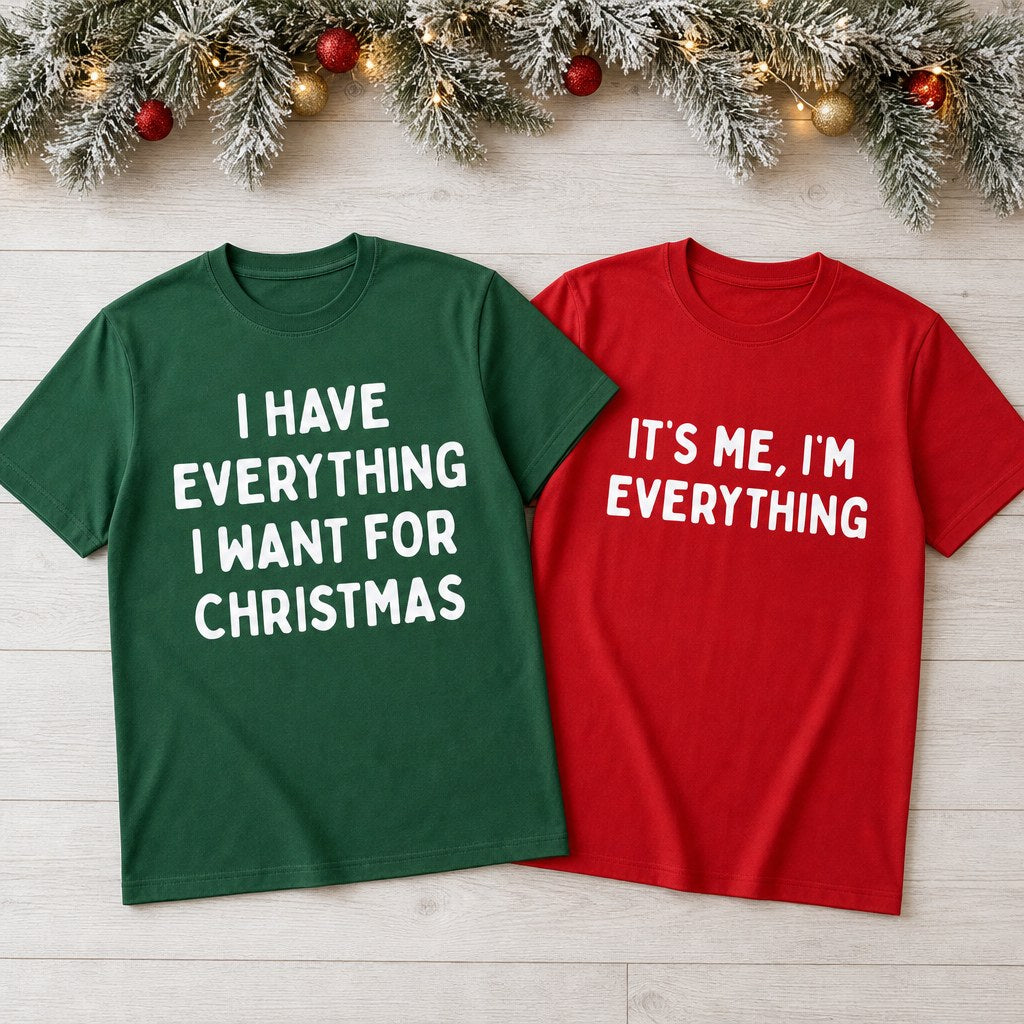 Both Christmas couples tees laid out on a wooden floor under a frosted pine garland
